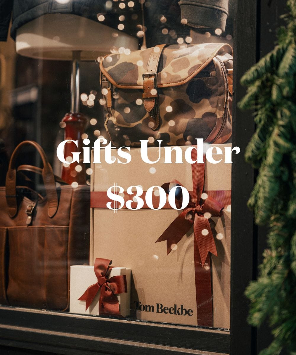 Store window display with gift boxes, a camo backpack, and a leather bag. Text reads: Gifts Under $300.