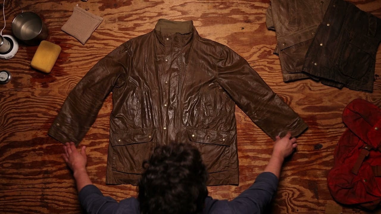 Person handling a brown waxed jacket on a wooden surface, surrounded by cleaning supplies and other jackets.