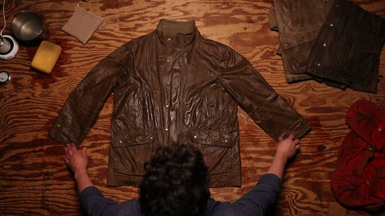 Person handling a brown waxed jacket on a wooden surface, surrounded by cleaning supplies and other jackets.