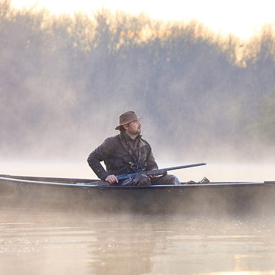 A person in a hat sits in a small boat on a misty lake with a fishing rod as trees are visible in the background.