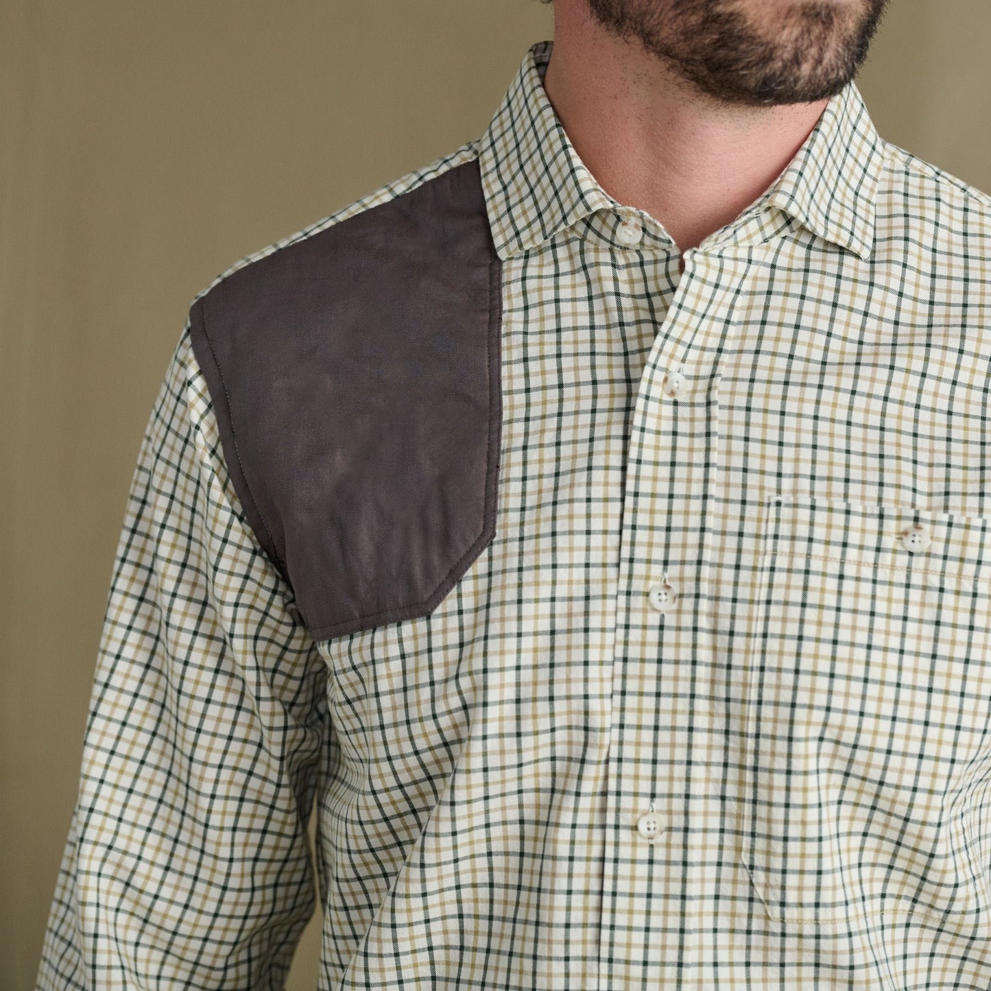 Man models Tom Beckbe Carrington Shooting Shirt with plaid pattern and dark patch, shown chest up on plain backdrop.