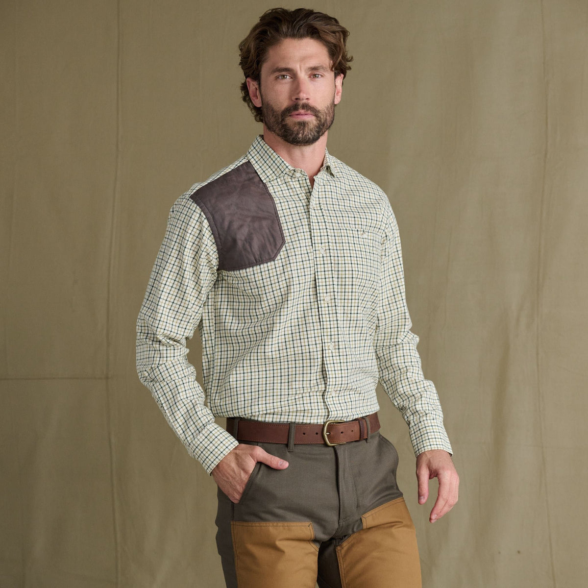 Man in Tom Beckbe Carrington Shooting Shirt with brown shoulder patch, two-tone pants, before plain backdrop.
