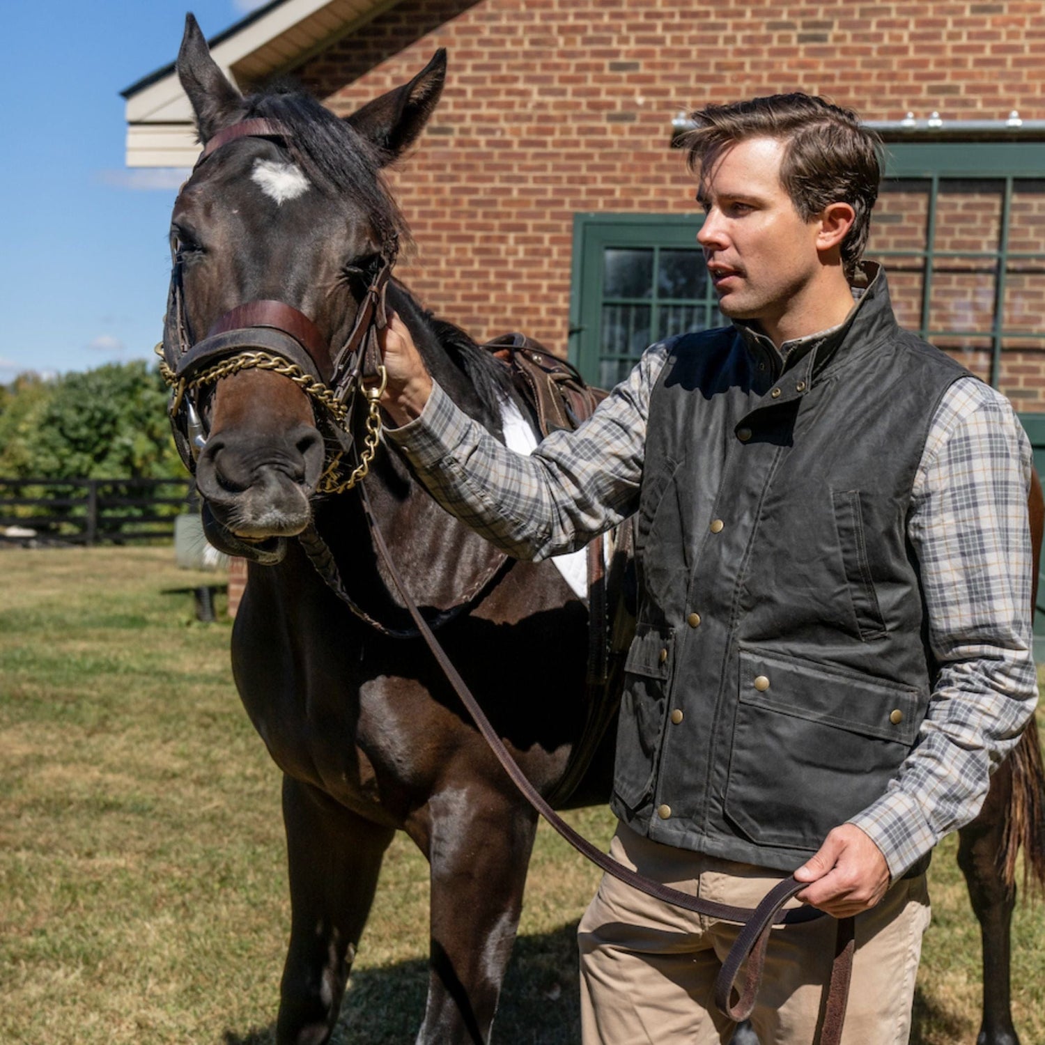 A man in a Tom Beckbe Kinsman Vest holds the reins of a brown horse outside a brick building on a sunny day.
