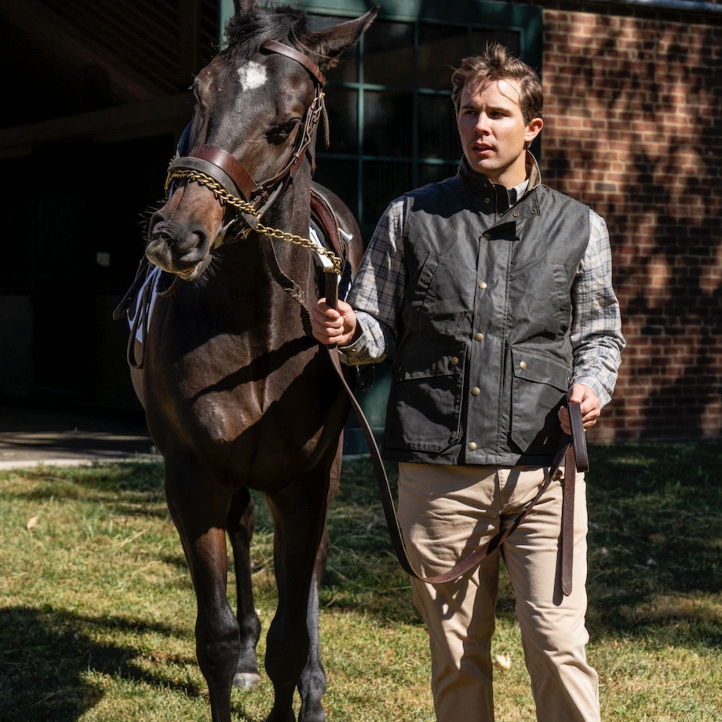 A person in a Tom Beckbe Kinsman Vest and plaid shirt guides a brown horse on grass near a brick building.