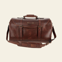 A brown leather duffel bag with a shoulder strap, handle, buckle closures, and brass hardware, set against a plain background.