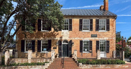 Historic two-story brick building with black shutters, arched doorway, and surrounding trees under a clear blue sky.