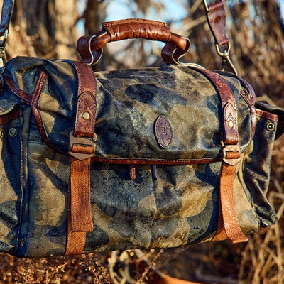 Camouflage-patterned duffel bag with brown leather straps and handle, outdoors on a natural background.
