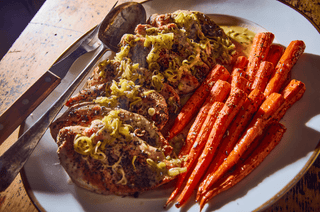 Plate of seasoned chicken topped with leeks, alongside roasted carrots, with a knife and spoon on a rustic wooden table.
