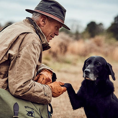 Man in a brown jacket and hat kneeling beside a black dog in a field. The dog has one paw raised, touching the mans arm.