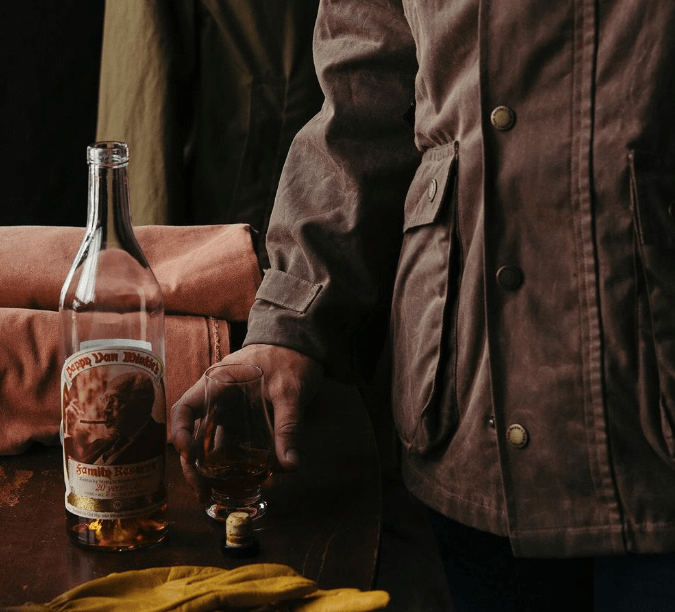 Person holding a whiskey glass next to a bottle on a table, with jackets and a pair of gloves nearby.