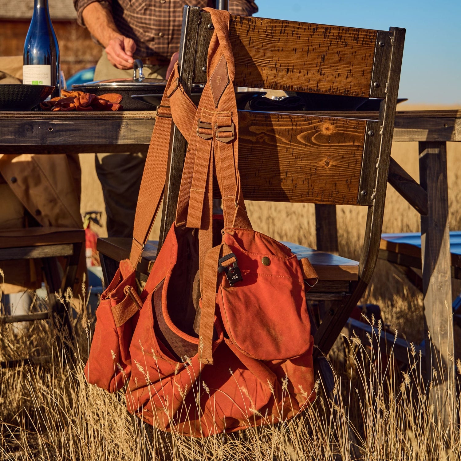 A Tom Beckbe Strap Vest hangs on a chair at a field table with a bottle and plate nearby.