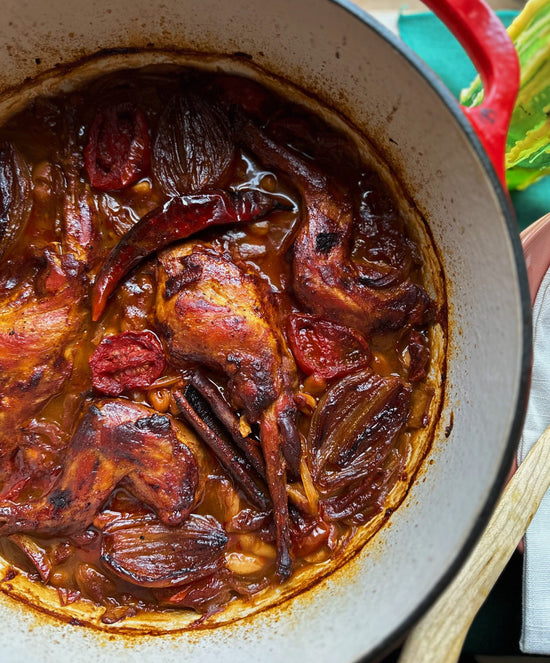 Cooked whole chicken with onions, tomatoes, and spices in a red Dutch oven, resting on a green and white surface.