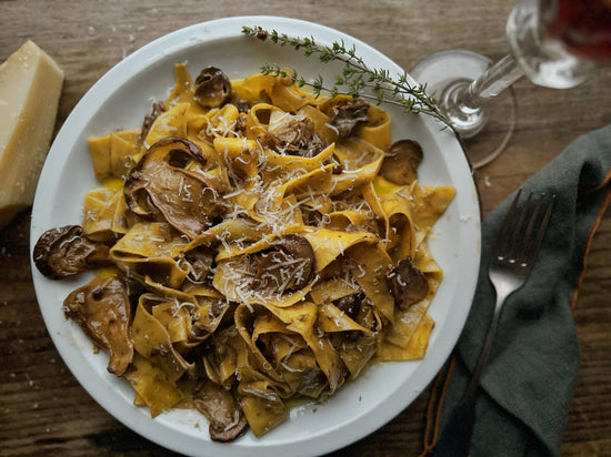 A plate of mushroom pappardelle pasta garnished with grated cheese and thyme, next to a glass of red wine on a wooden table.