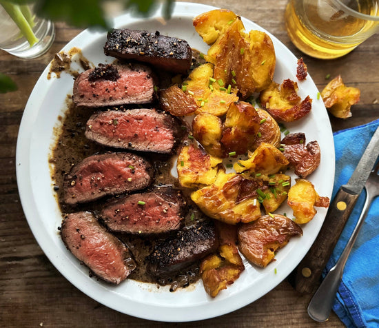 Plate of sliced steak seasoned with pepper and salt served with crispy, smashed potatoes and a drink on a wooden table.
