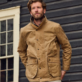 A man in a Tom Beckbe Tensaw Jacket stands against a dark wooden wall and window, showcasing its durable style.