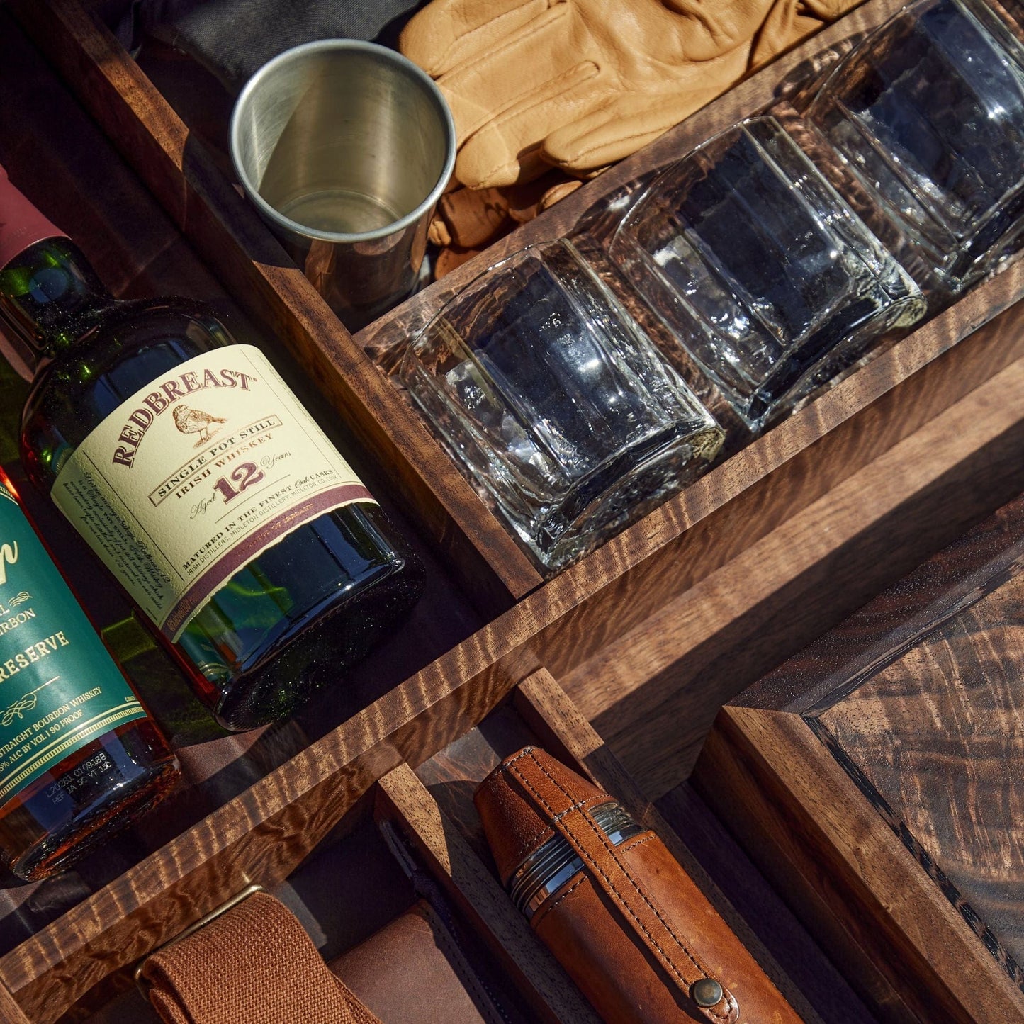 A wooden box with whiskey bottles, Terrane No. 12 Glass by Tom Beckbe, a metal cup, gloves, and leather accessories.