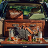 Car trunk setup with wooden shelves, bags, bottles, Tom Beckbes Terrane No. 12 Glasses, and an ice bucket outdoors.