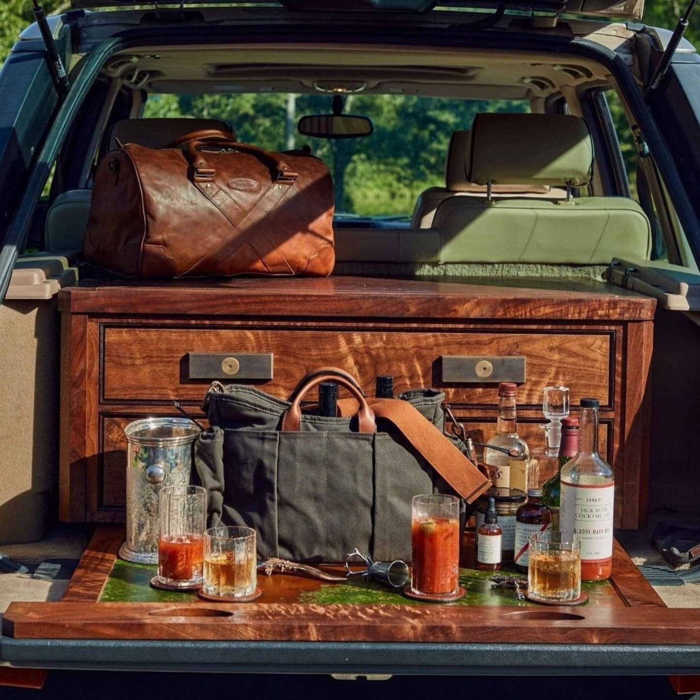 Car trunk setup with wooden shelves, bags, bottles, Tom Beckbes Terrane No. 12 Glasses, and an ice bucket outdoors.