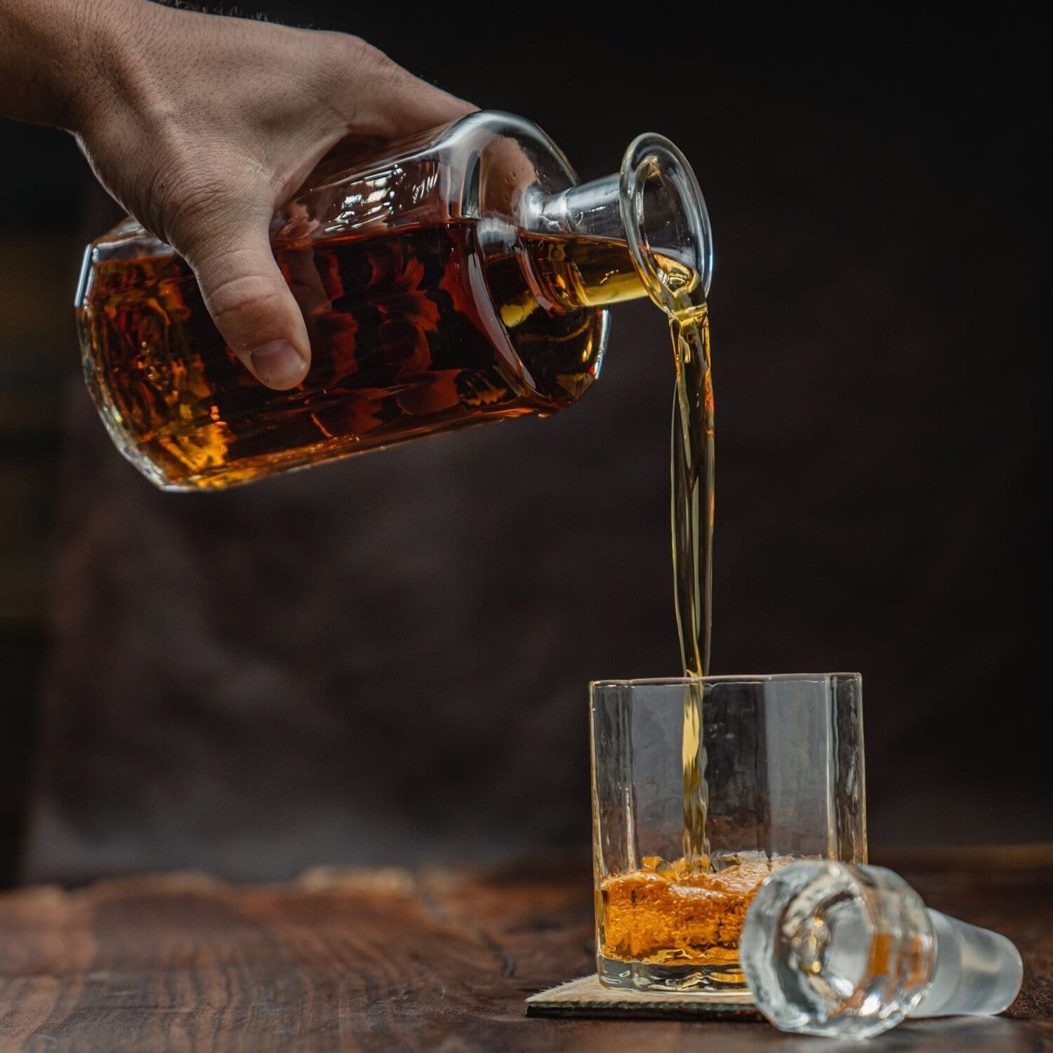 A hand pours amber liquid from a decanter into Terrane No. 12 Glasses by Tom Beckbe on wood, stopper nearby.