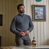 A man in a Tom Beckbe Albemarle Henley Sweater stands indoors near a table, with art on a cream-paneled wall behind him.