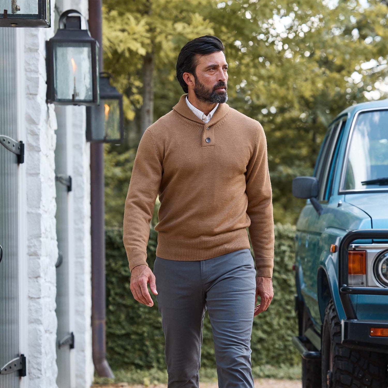 Man in Tom Beckbe Henry Shawl Sweater in Royal Alpaca and gray pants walks outside by a white building and blue vehicle.