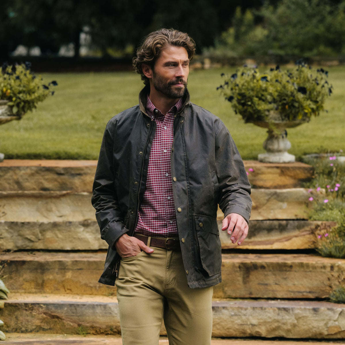 Man in a Tom Beckbe Baldwin Jacket, checked shirt, and khakis walks outdoors near stone steps and greenery.