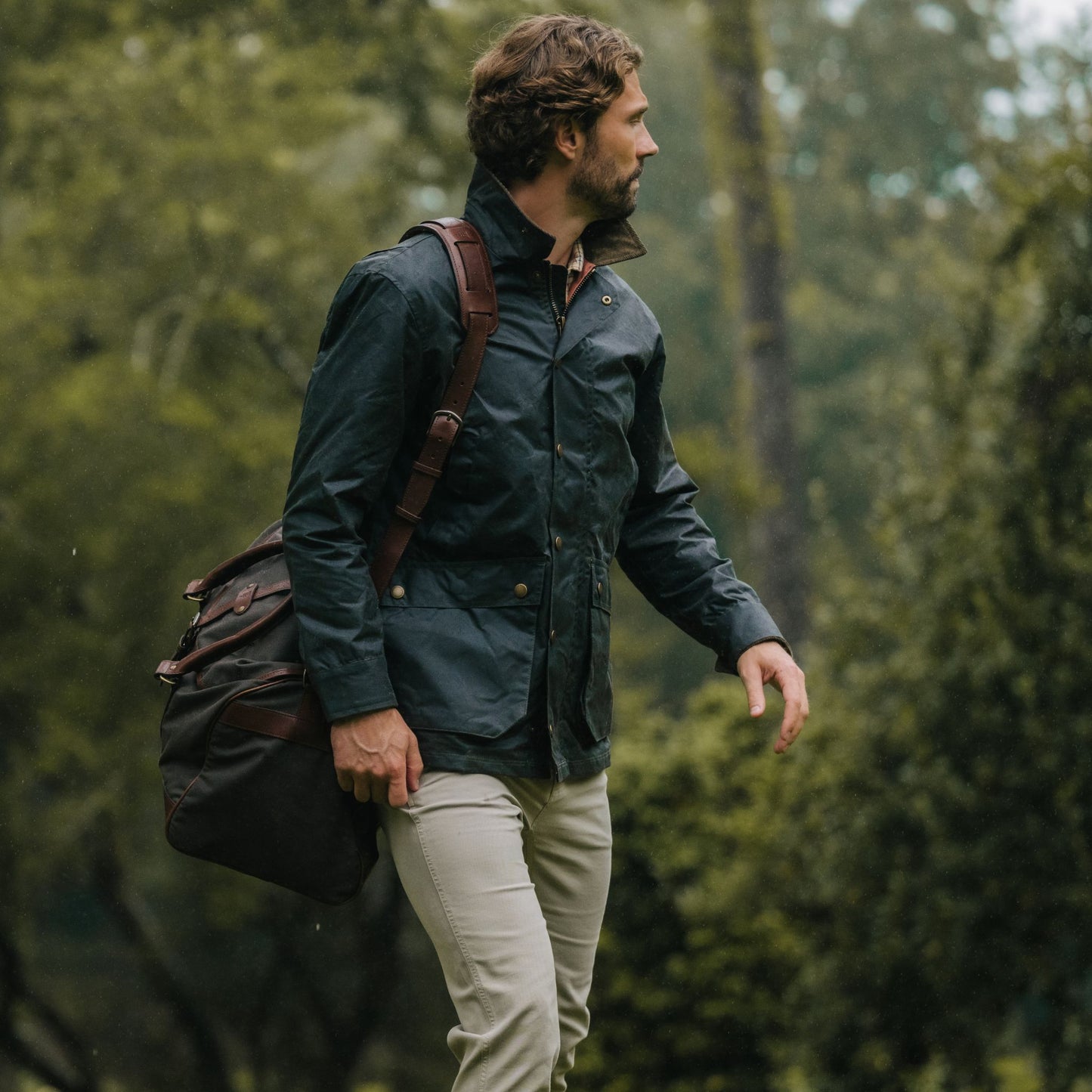 Man in a Tom Beckbe Baldwin Jacket with a brown shoulder bag walks outdoors in a green, wooded area.