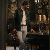 A man in a Tom Beckbe Baldwin Jacket and khakis stands indoors, holding a brown leather bag by the table.
