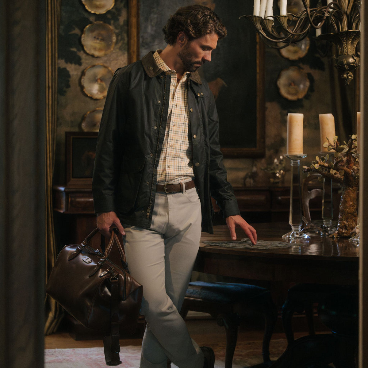 A man in a Tom Beckbe Baldwin Jacket and khakis stands indoors, holding a brown leather bag by the table.
