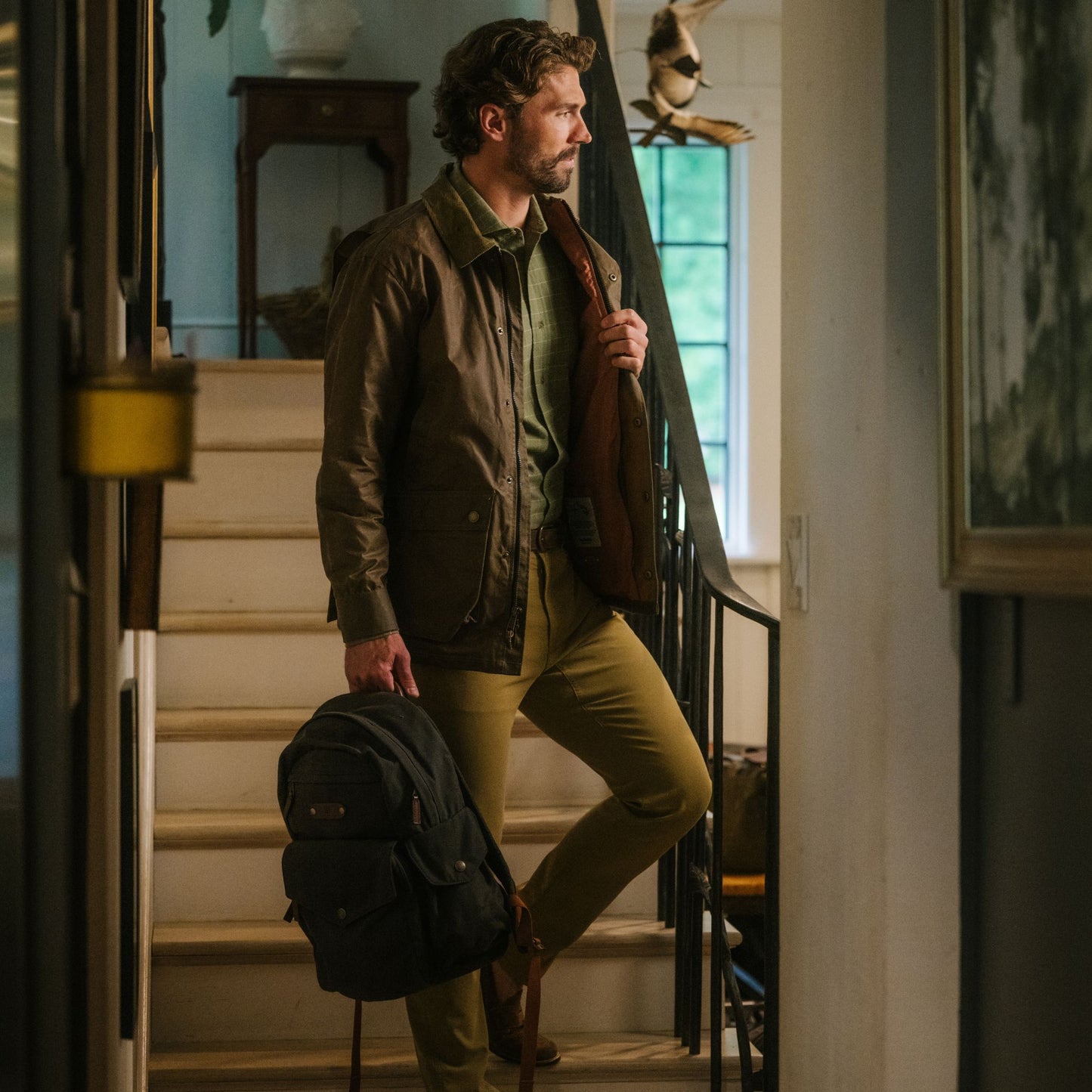 Man stands indoors on a staircase, holding a backpack and his Tom Beckbe Baldwin Jacket over his shoulder.