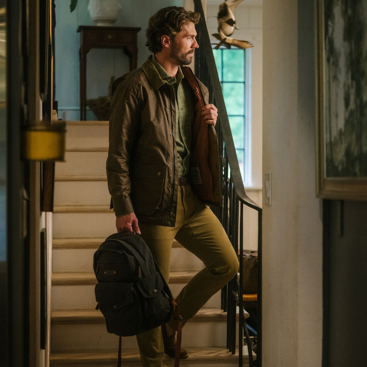 Man stands indoors on a staircase, holding a backpack and his Tom Beckbe Baldwin Jacket over his shoulder.