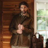 Bearded man in Tom Beckbe’s Baldwin Jacket stands indoors by a window next to leather bags on a bench.