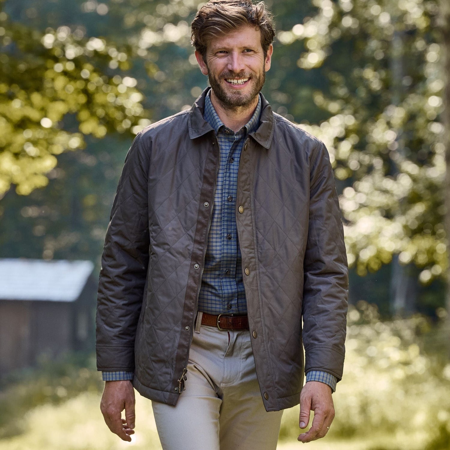 Man outdoors in a checked shirt and Tom Beckbe Braddock Quilted Jacket, smiling with trees and a wooden structure behind.