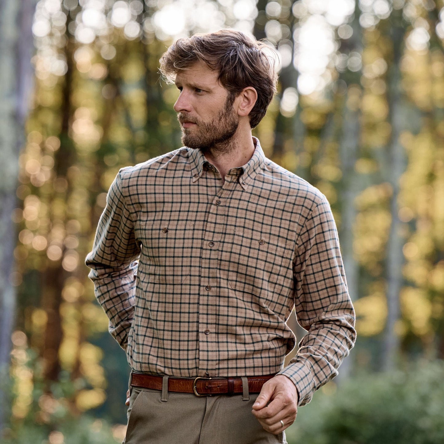 Man in a Tom Beckbe Dixon Brushed Cotton Twill Shirt and brown belt stands outdoors in a wooded area, looking aside.