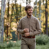 A man in a Tom Beckbe Dixon Brushed Cotton Twill Shirt and khaki pants smiles outdoors in a sunlit forest clearing.