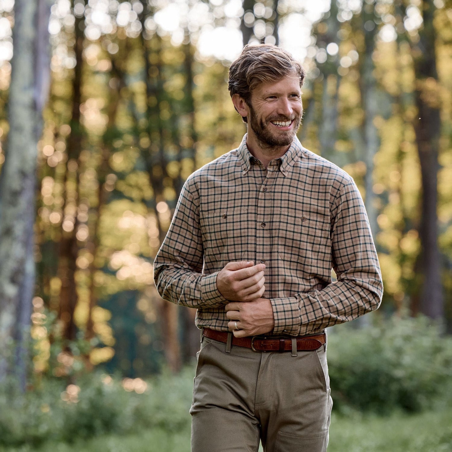 A man in a Tom Beckbe Dixon Brushed Cotton Twill Shirt and khaki pants smiles outdoors in a sunlit forest clearing.