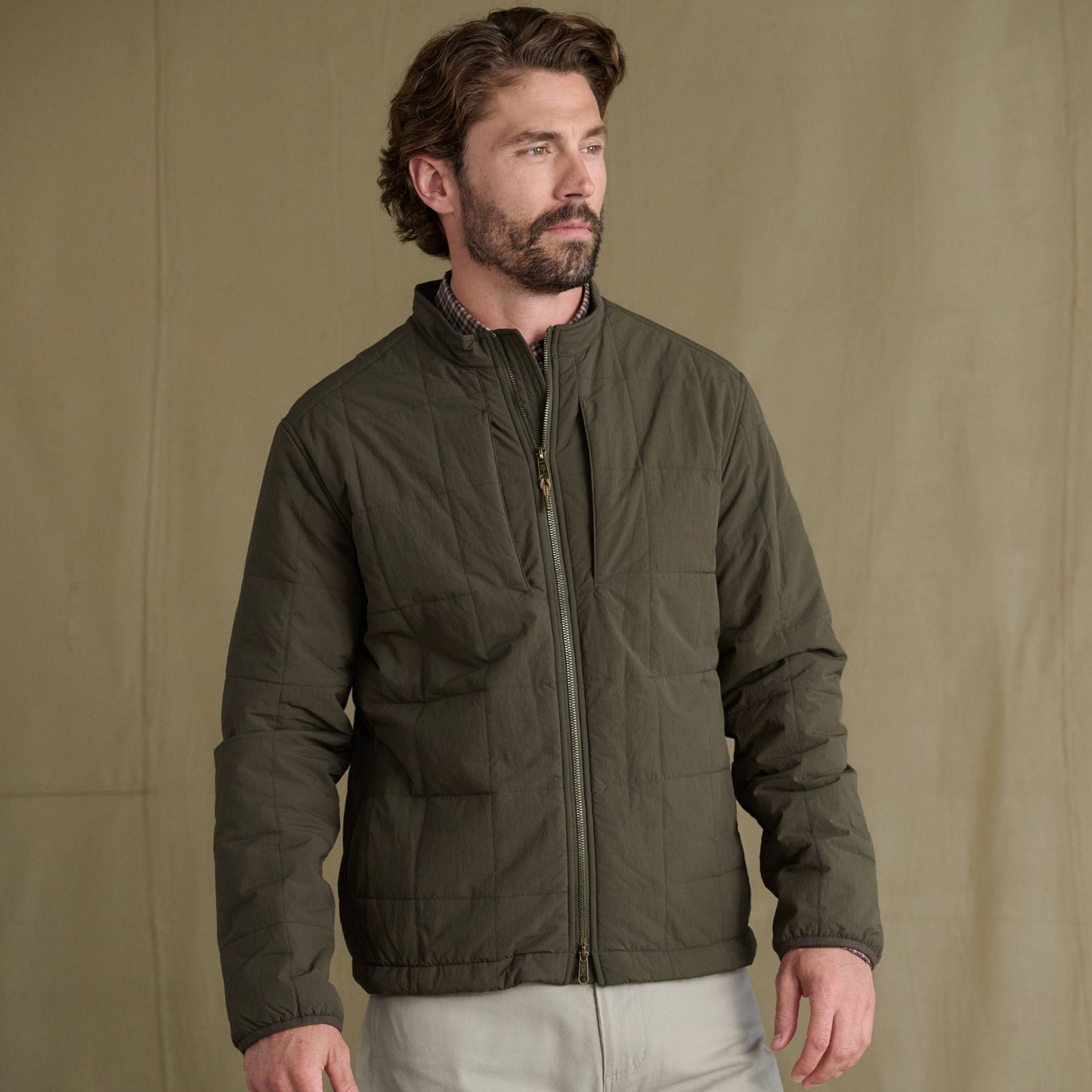Man wears Tom Beckbe Burgess Quilted Jacket and light pants, standing against a plain tan background.