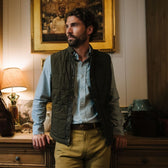 Man in Burgess Quilted Vest by Tom Beckbe stands by dresser with art, lamp, and pottery in a warmly lit room.