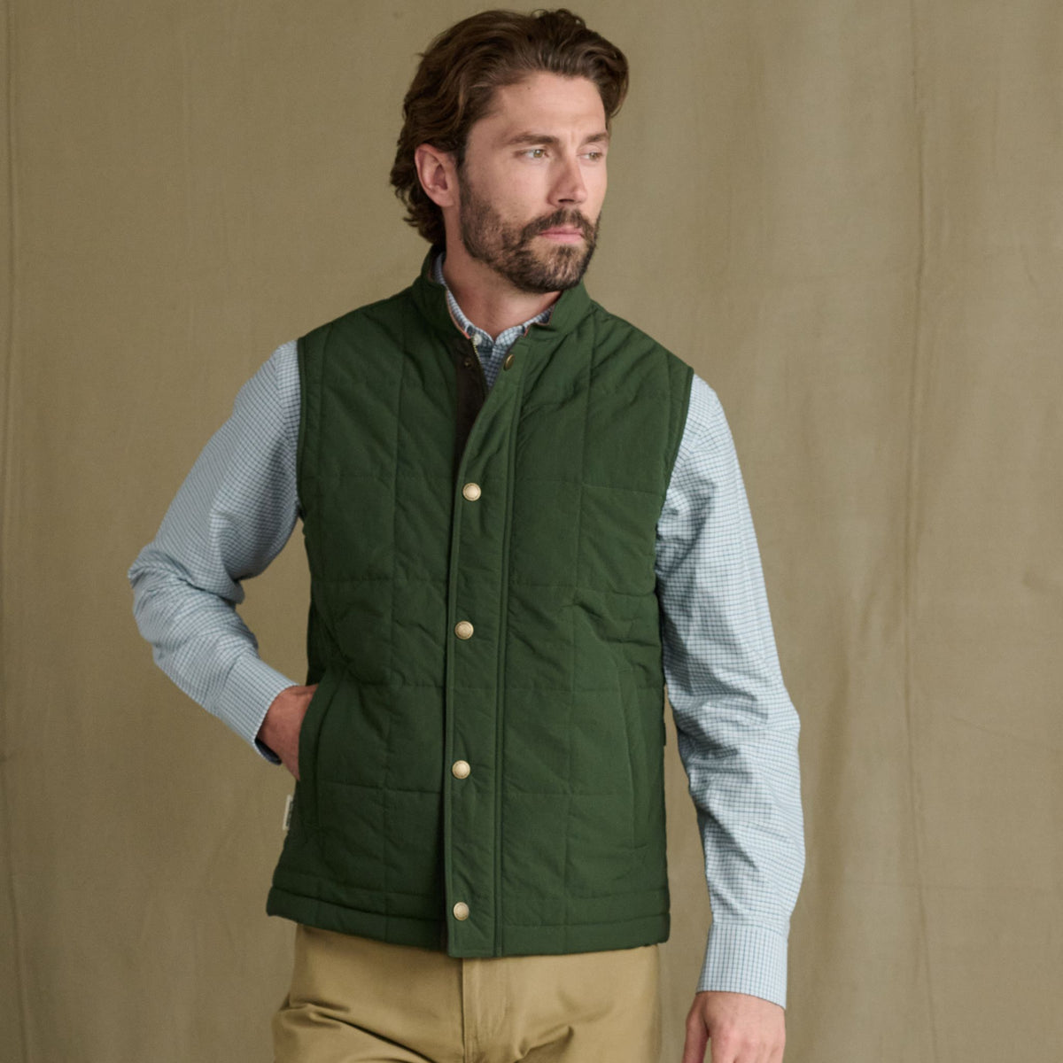 Man in Tom Beckbe Burgess Quilted Vest over light blue check shirt and khakis, standing by beige background.