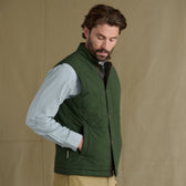 Man wears Tom Beckbe’s Burgess Quilted Vest over a blue checkered shirt against a beige background.