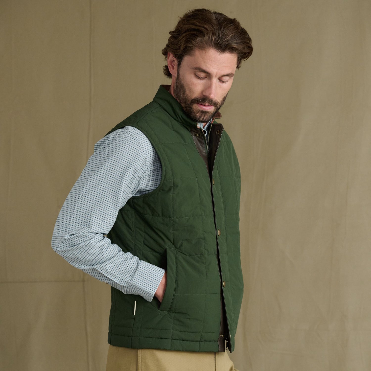 Man wears Tom Beckbe’s Burgess Quilted Vest over a blue checkered shirt against a beige background.