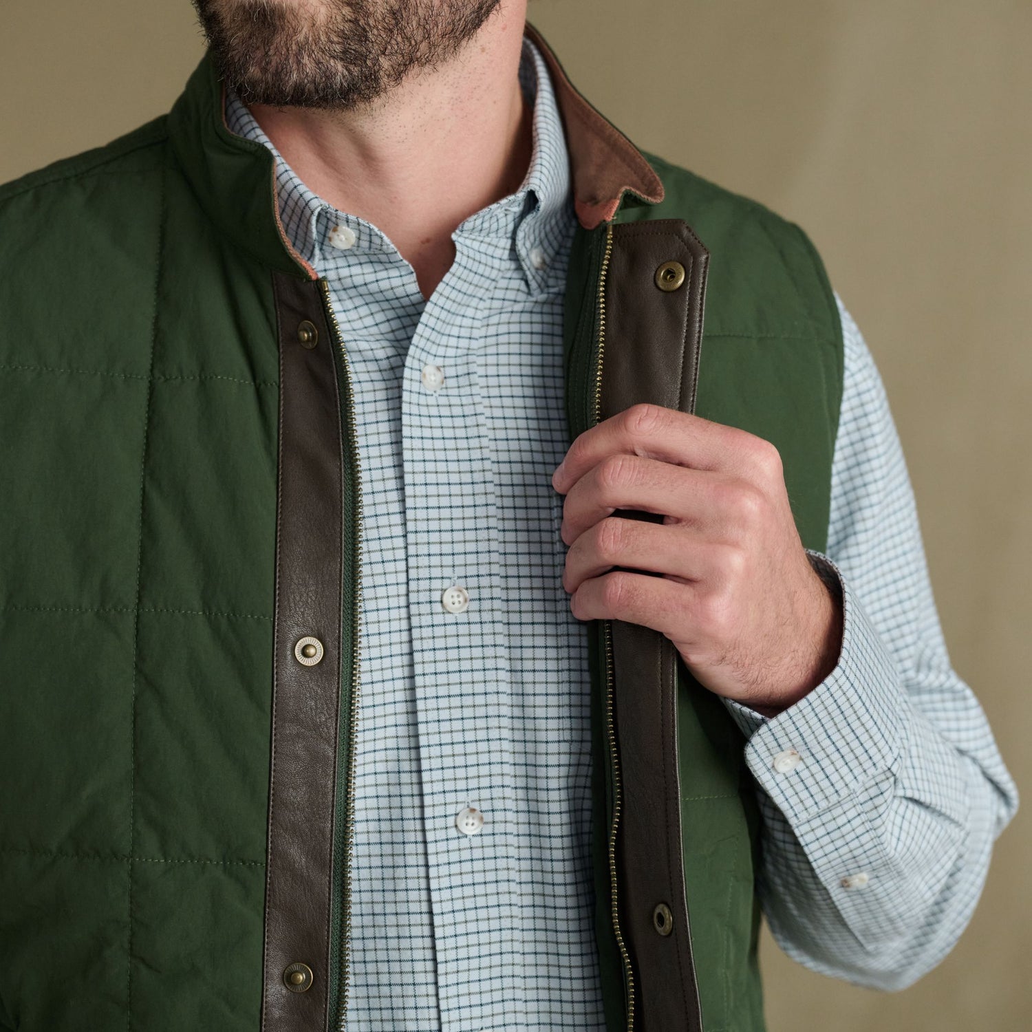 Man in Tom Beckbe Burgess Quilted Vest over a blue checkered shirt, holding the open vest near its zipper with one hand.