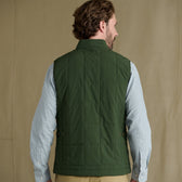 Man wears Tom Beckbe Burgess Quilted Vest in green over a light blue check shirt, facing away from the camera.