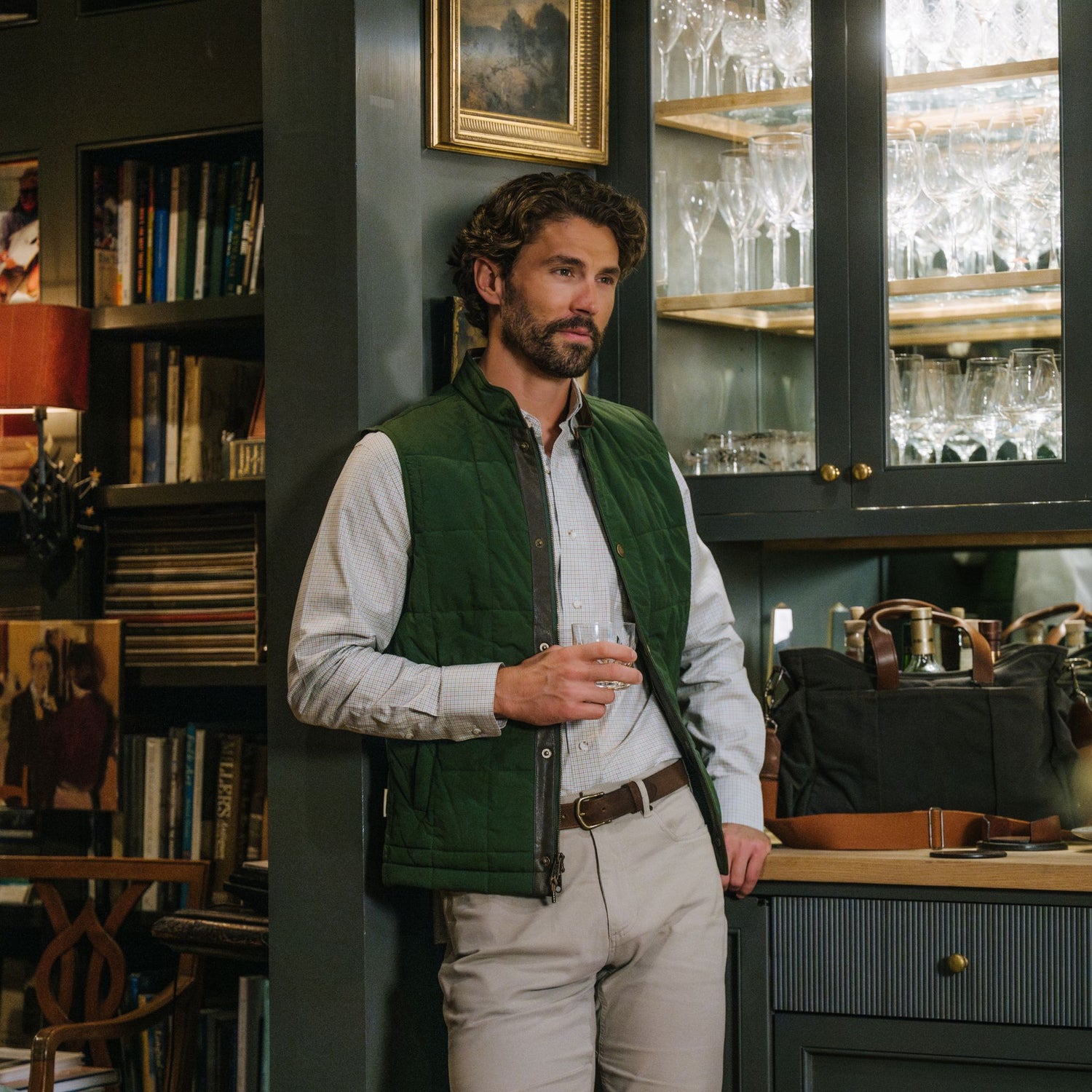 Man in Tom Beckbe Burgess Quilted Vest holds a glass by a glassware cabinet, surrounded by bookshelves and art.