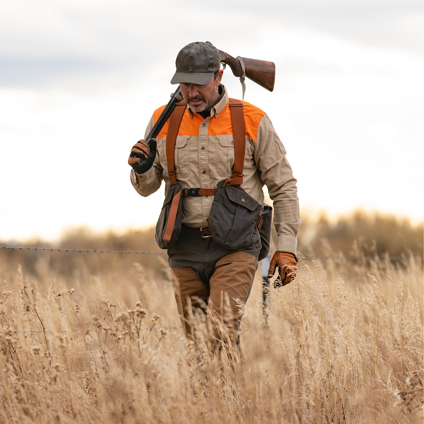 Man in Tom Beckbe Burwell Brush Pant with shotgun over shoulder walks through tall dry grass in open field.
