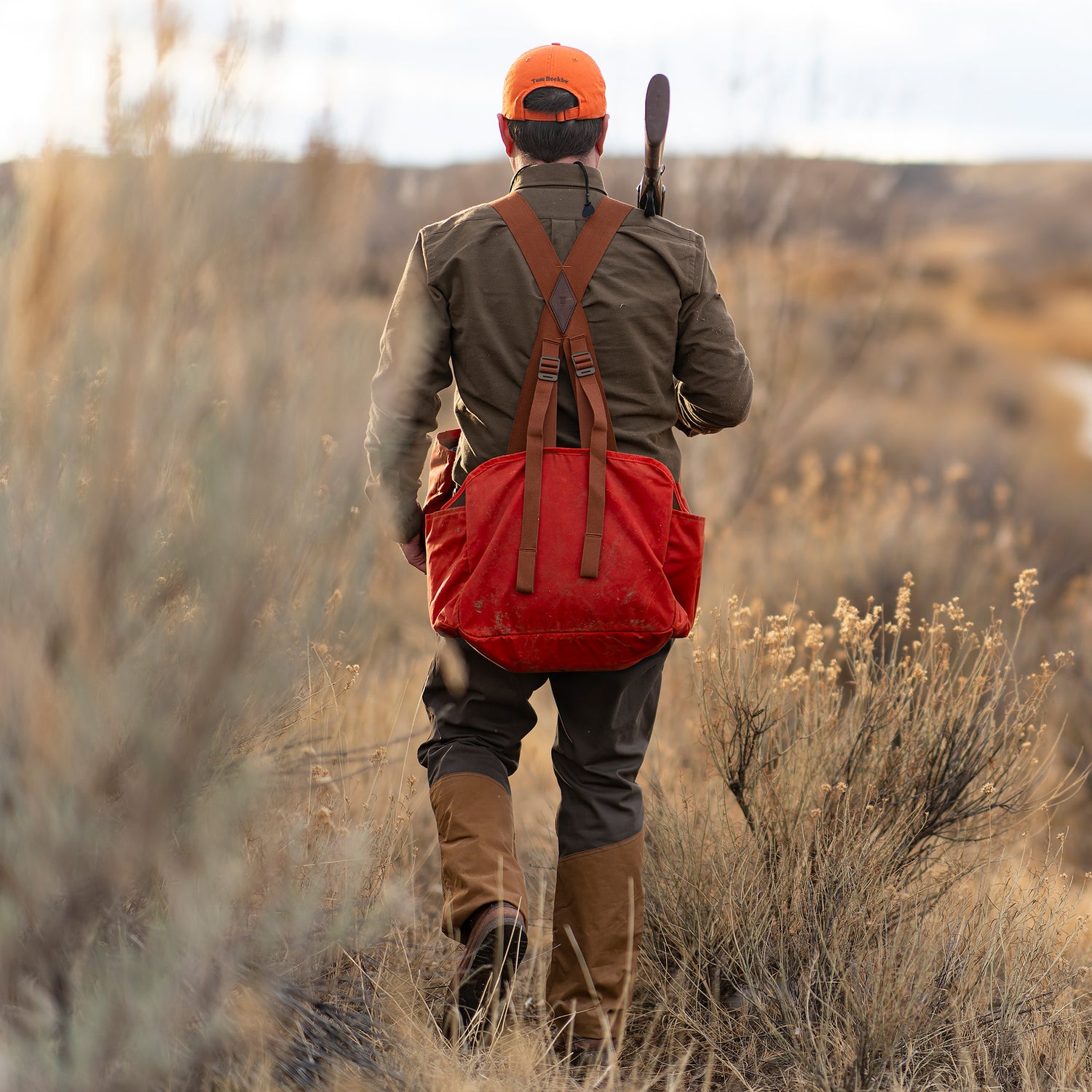 Wearing Tom Beckbe Burwell Brush Pant and an orange cap, a person walks through dry grass with a red bag and rifle.