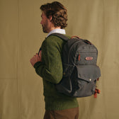Man in a green sweater wears a Tom Beckbe Canvas Backpack, standing against a plain tan background.