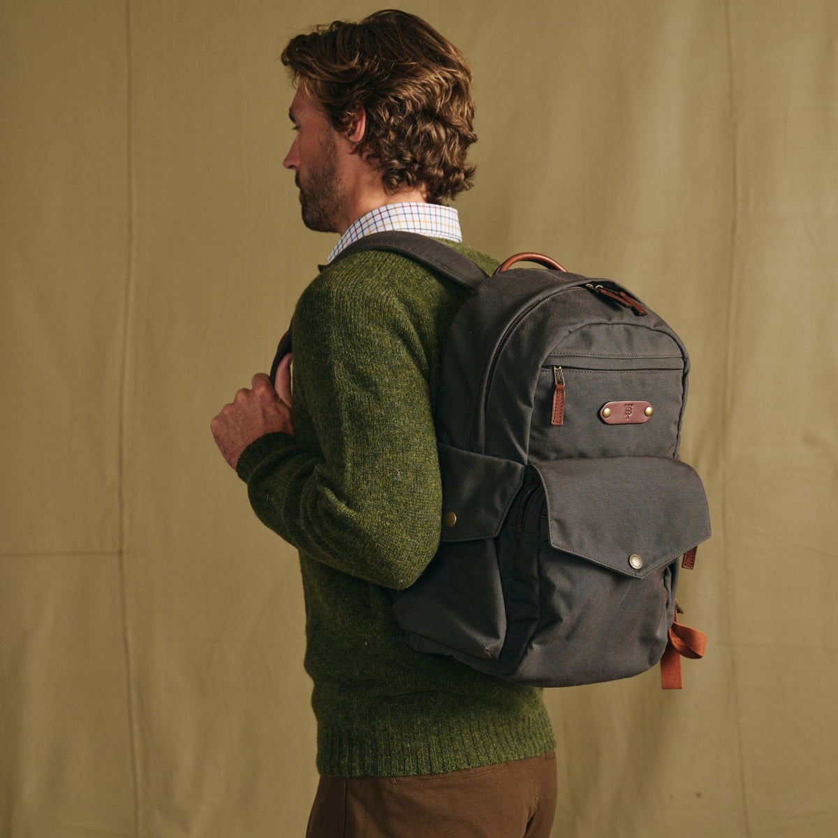 Man in a green sweater wears a Tom Beckbe Canvas Backpack, standing against a plain tan background.