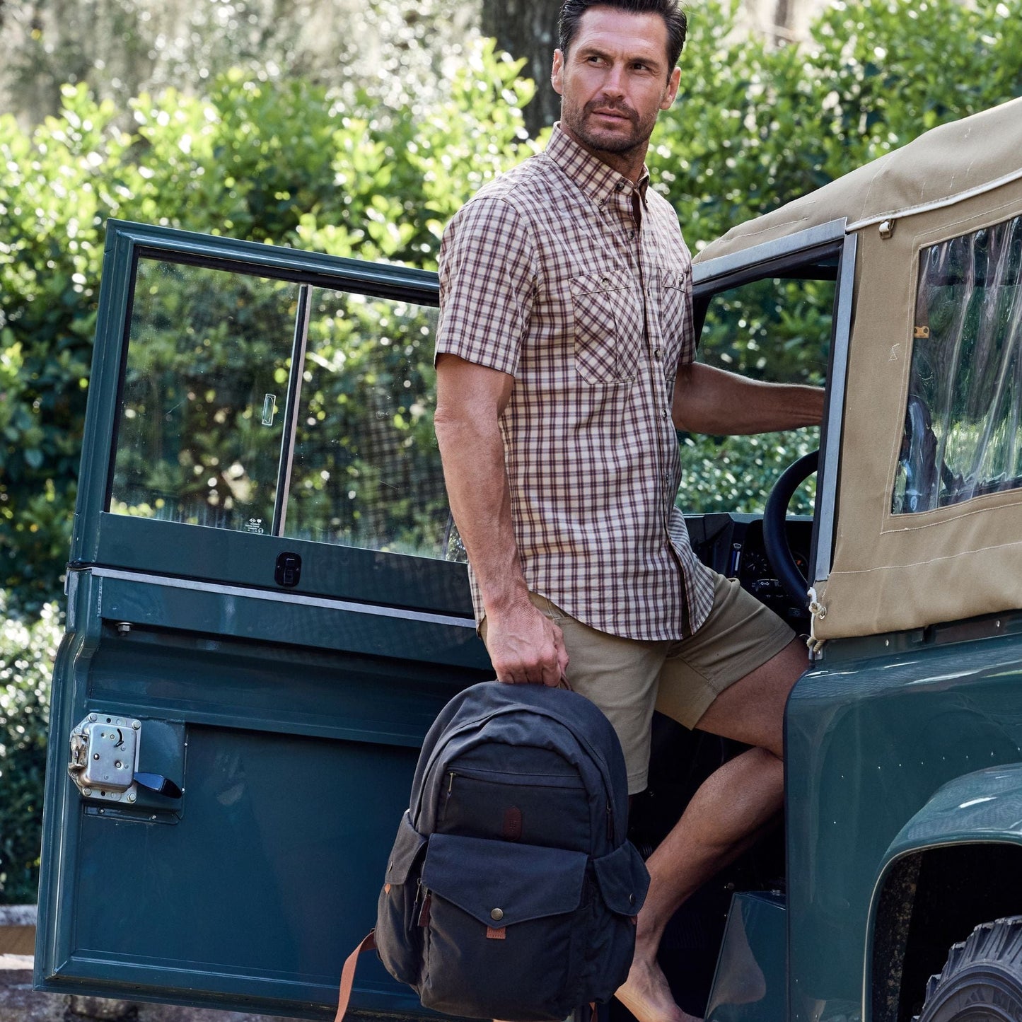 A man with a Tom Beckbe Canvas Backpack exits an off-road vehicle, surrounded by lush greenery.
