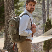 Man in a light shirt and khaki pants stands near trees with a Tom Beckbe Canvas Backpack featuring a laptop sleeve.